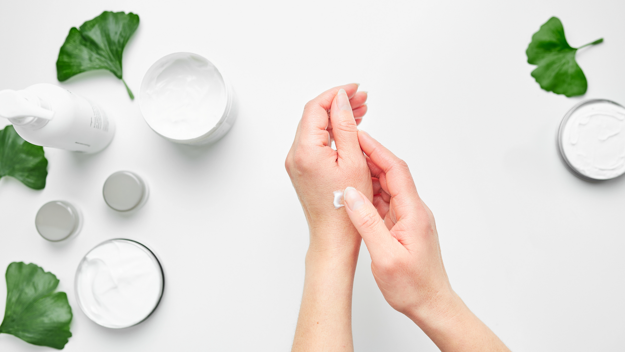 Person putting hand cream on hands. Cream containers and green leafs.