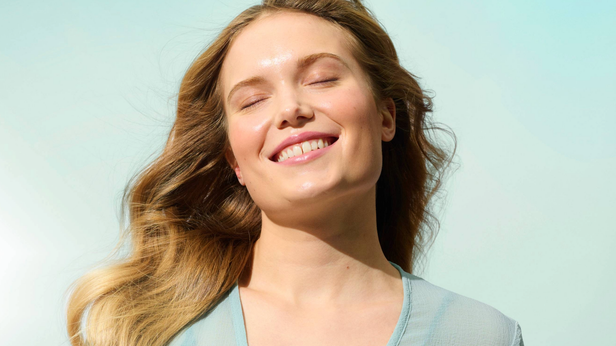 Pitkähiuksinen, hymyilevä nainen pitää silmiään kiinni vaaleansininen tausta