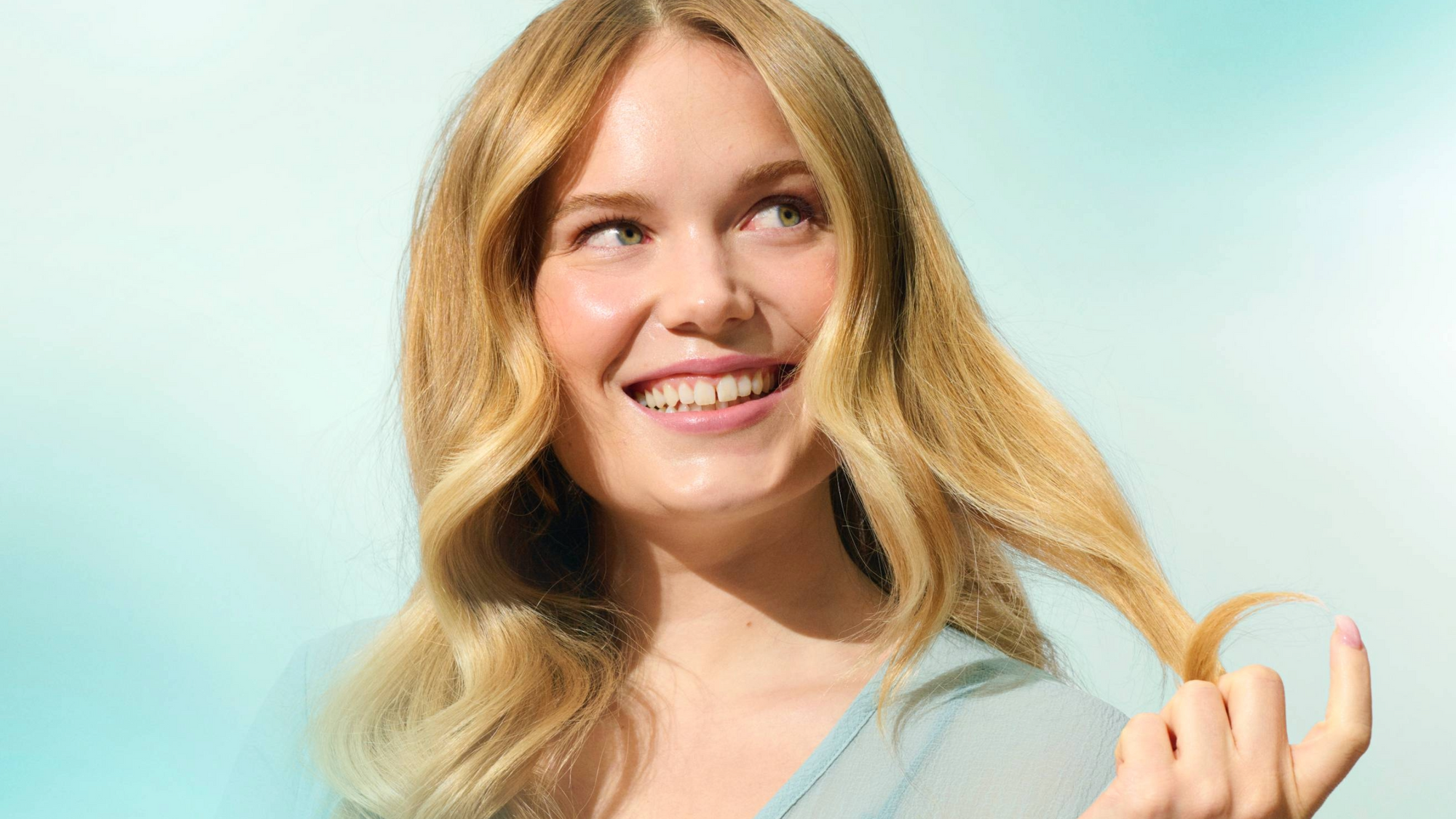 Pitkä- ja vaaleahiuksinen, hymyilevä nainen vaalean sininen tausta